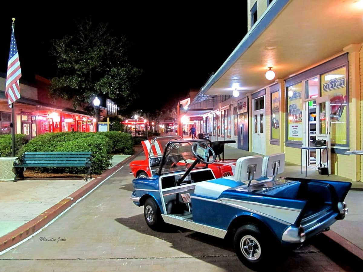Oldtimer entlang einer hell erleuchteten Straße in der Old Town Kissimmee geparkt, mit Neon-Schaufenstern, Bänken und einer amerikanischen Flagge bei Nacht.