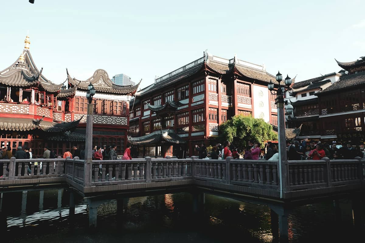 Lebhafte Szene in Shanghais Altstadt (Nanshi) mit traditionellen chinesischen Gebäuden, roten Laternen und einer beliebten Fußgängerbrücke über Wasser an einem hellen Tag.