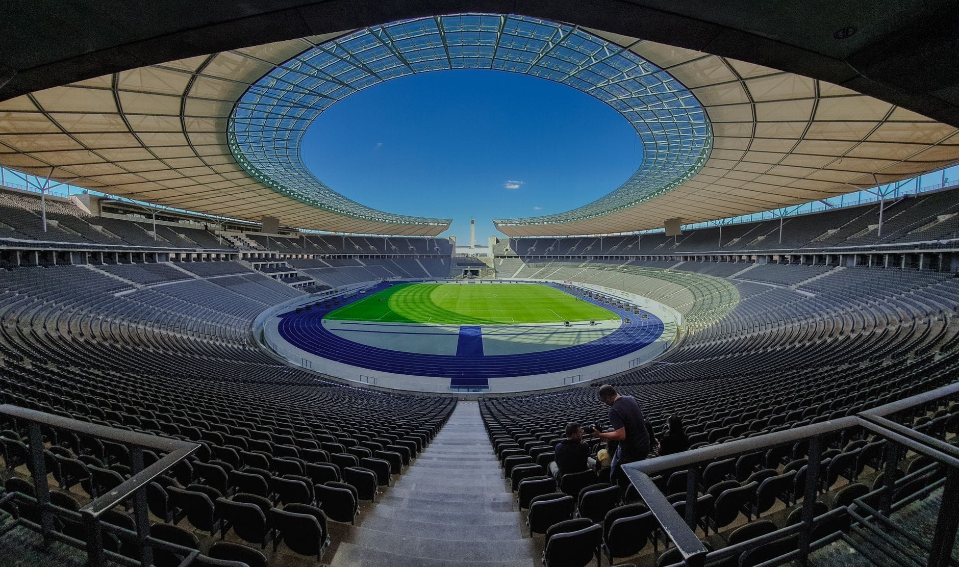 Vista grandangolare dagli spalti dell'Olympiastadion Berlin: il moderno tetto ovale, la pista blu, il campo verde e la curva continua dei seggiolini vuoti sotto un cielo sereno.