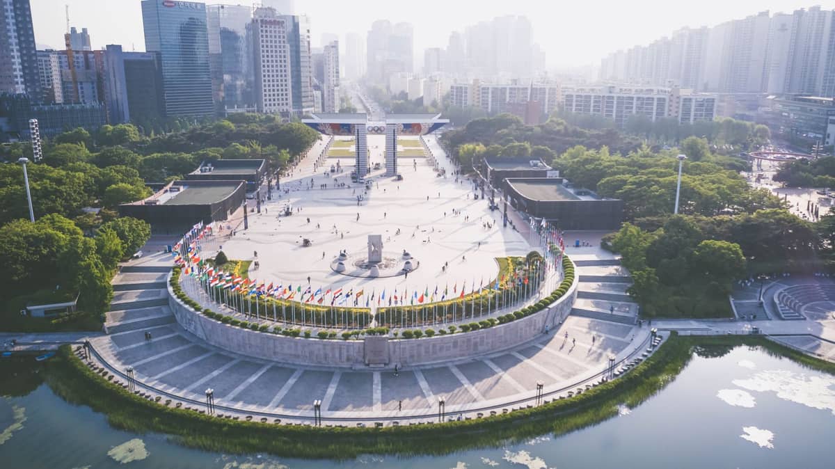 Vista aérea do Parque Olímpico de Seul mostrando a Peace Plaza, o display de bandeiras, o simbólico Portão da Paz Mundial e os prédios ao redor durante um dia ensolarado.