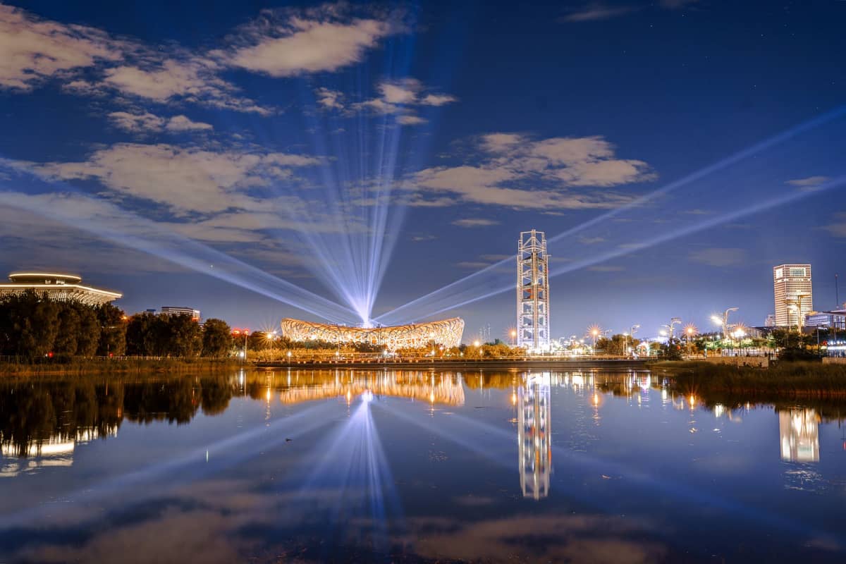 Nachtansicht des Olympiaparks Peking mit dem beleuchteten Vogelnest-Stadion, dem Olympischen Turm und Lichtstrahlen, die sich in einem ruhigen See spiegeln.