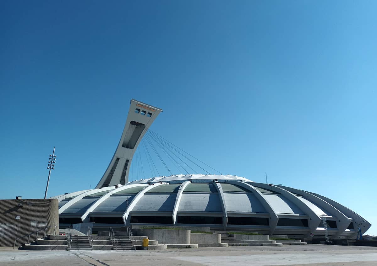 Weitläufige Landschaftsansicht des Olympiastadions Montreal mit dem markanten geneigten Turm vor einem klaren blauen Himmel, mit Blick auf den Haupteingang und die architektonischen Details.