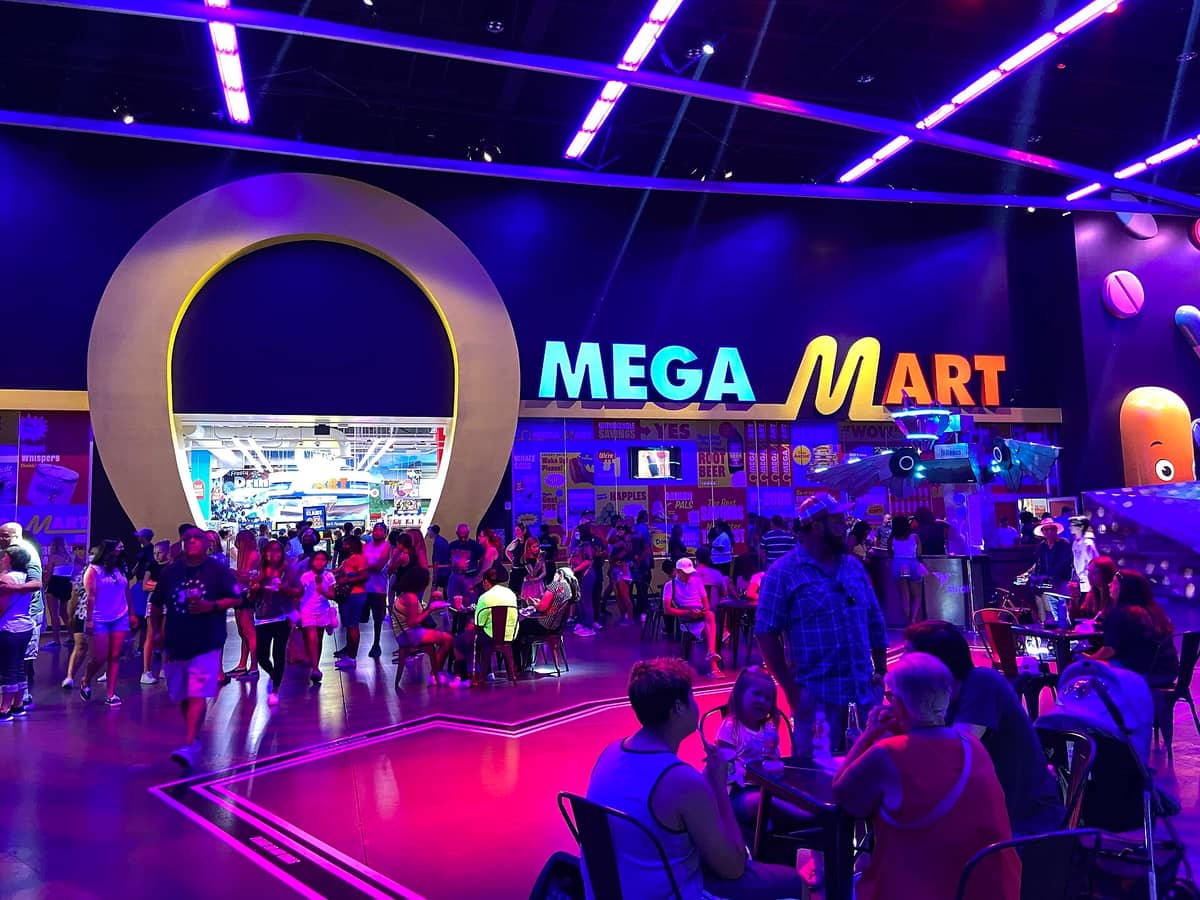 Kerumunan di pintu masuk Omega Mart di AREA15 Las Vegas yang penuh warna neon, dengan papan OMEGA MART besar di atasnya.