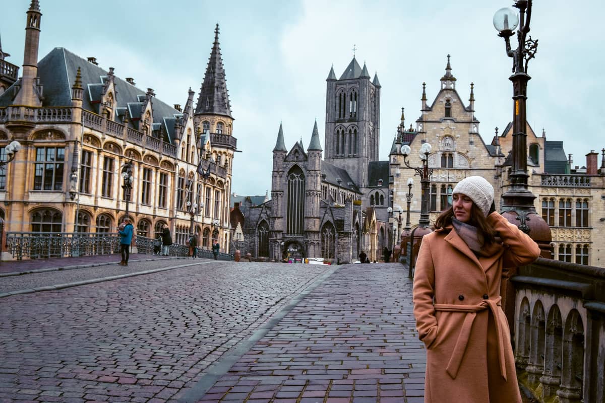 View of Ghent's medieval city center with St. Nicholas' Church and historic buildings, cobblestone street, and pedestrians, taken on an overcast morning