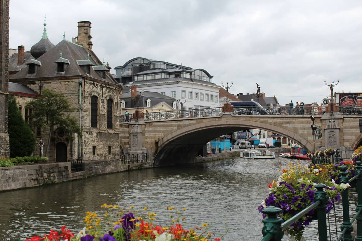 View of St. Michael's Bridge over a canal in Ghent, with historic stone buildings, flowers in the foreground, and boats under the bridge.
