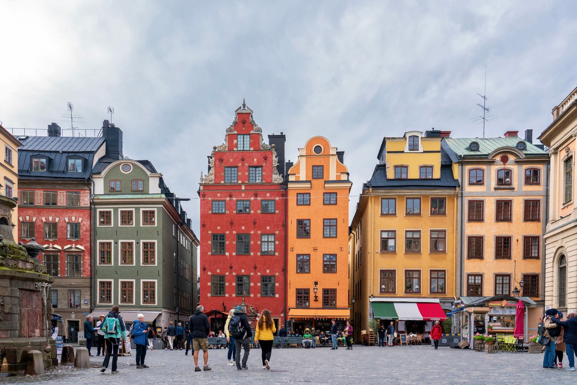 ด้านหน้าอาคารประวัติศาสตร์หลากสีสันและจัตุรัสหลักของ Gamla Stan ในสตอกโฮล์ม พร้อมผู้คนเดินเที่ยว ร้านค้าและคาเฟ่ที่เปิดในยามเช้า