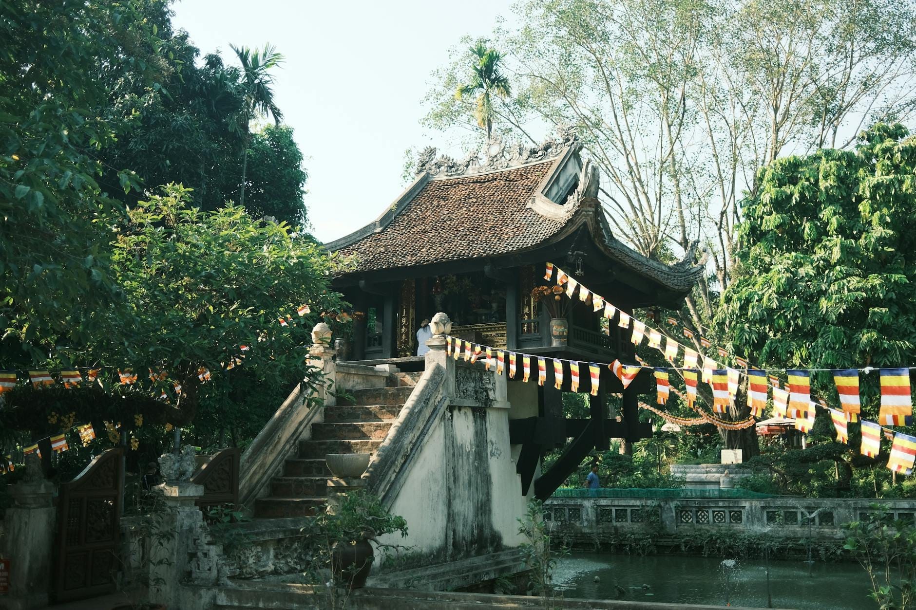 The One Pillar Pagoda, a small wooden Buddhist shrine perched on a single stone pillar in the middle of a lotus pond, with trees and colorful Buddhist flags around it in Hanoi, Vietnam.