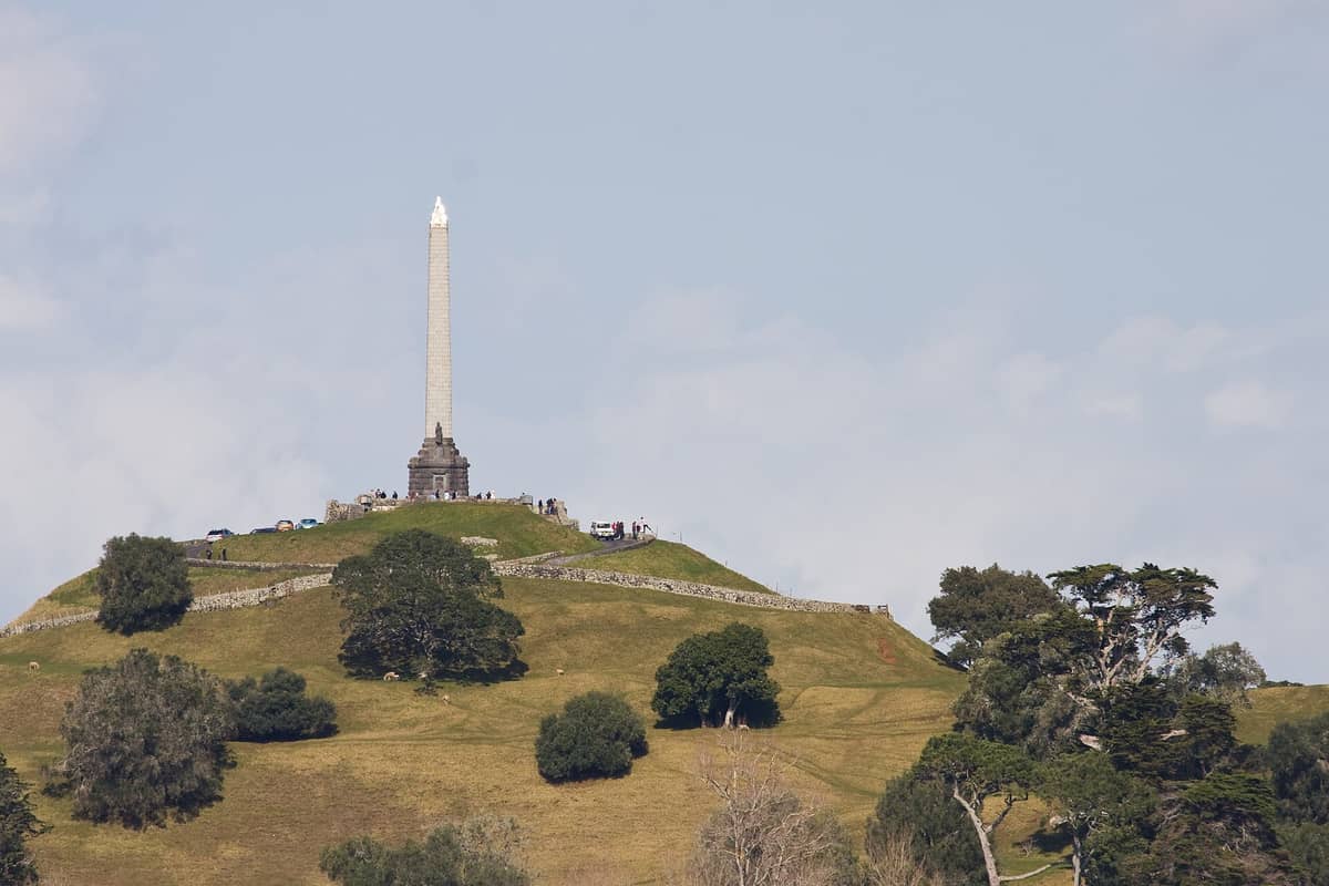 Nahaufnahme des Gipfels von One Tree Hill (Maungakiekie) in Auckland mit dem ikonischen Obelisken und den grasbedeckten Vulkanhängen unter blauem Himmel.