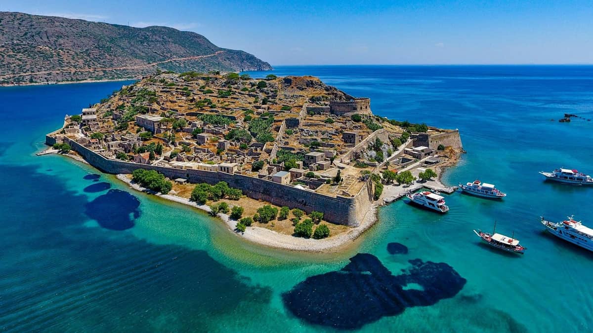 Aerial view of Spinalonga island with Venetian fortifications, turquoise waters, and tour boats anchored nearby.
