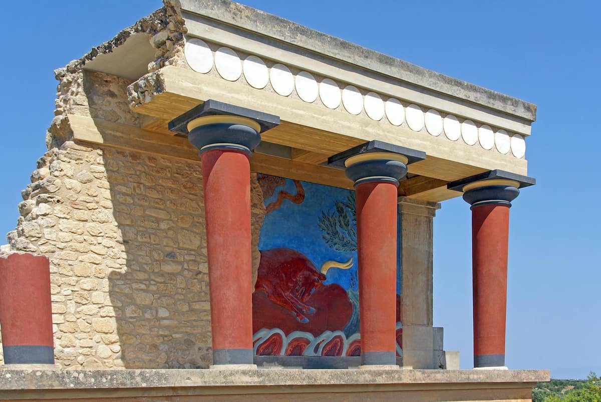 Ruins of Knossos Palace near Heraklion, featuring red Minoan columns, stone walls, and colorful fresco under a clear blue sky.