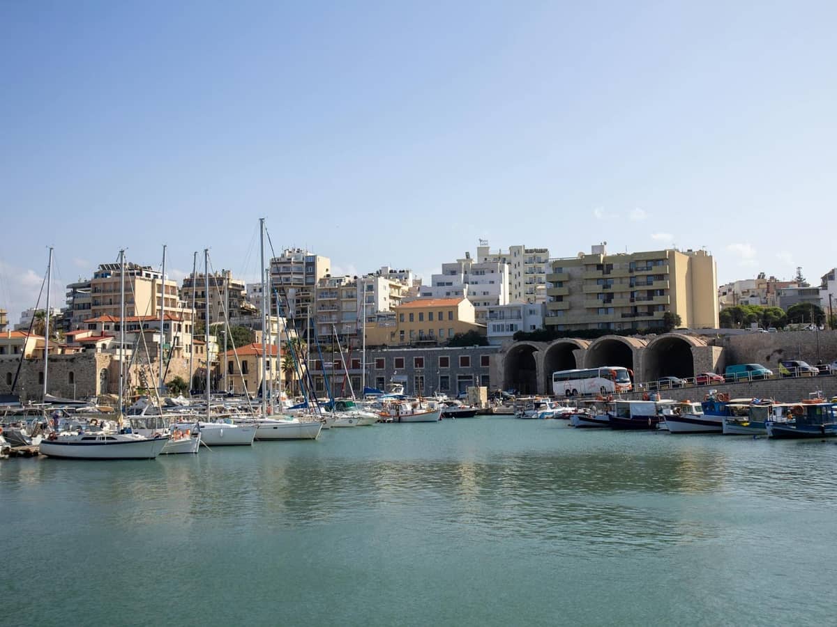 Yachts and fishing boats moored in the harbor at Heraklion, with the city and waterfront buildings in the background under a clear sky.