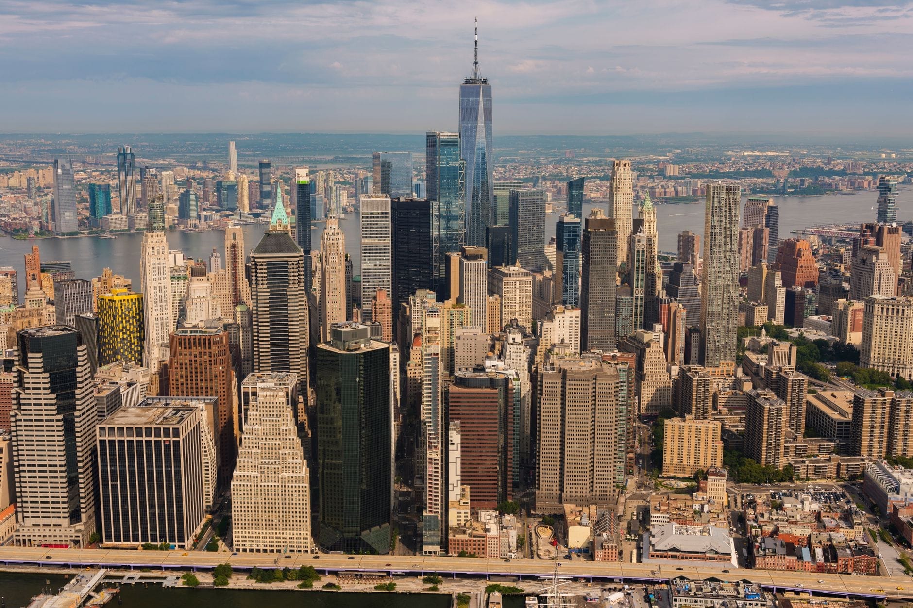 Vista aérea deslumbrante dos arranha-céus do Baixo Manhattan, incluindo o One World Trade Center, com o Rio Hudson ao fundo sob um céu parcialmente nublado.