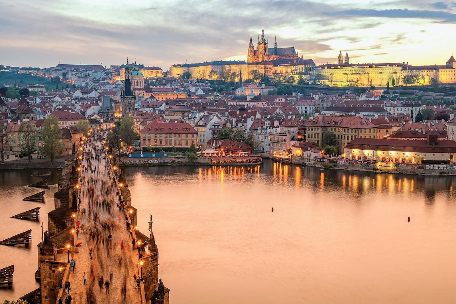 Luftaufnahme der Karlsbrücke und der Prager Burg bei Sonnenuntergang, mit der Moldau, die die Stadtlichter spiegelt, und Menschen auf der Brücke.