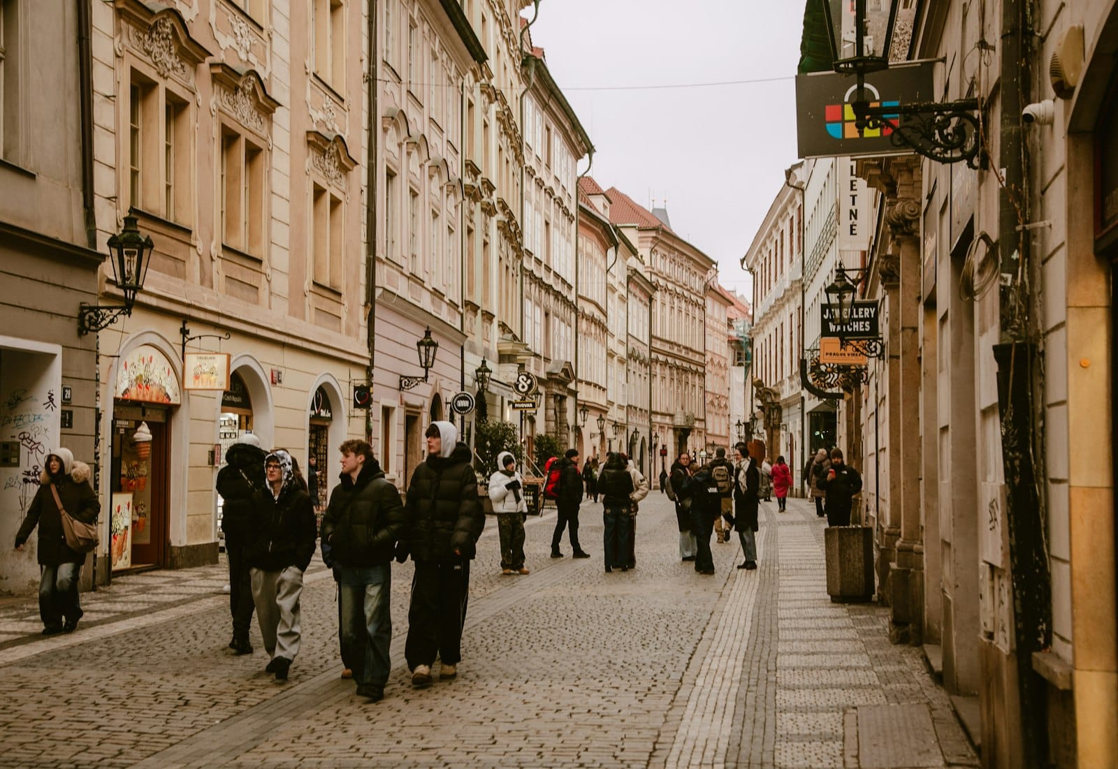 Menschen, die durch eine ruhige, charmante Kopfsteinpflasterstraße mit historischen Gebäuden und kleinen Geschäften in einem Prager Stadtviertel spazieren.