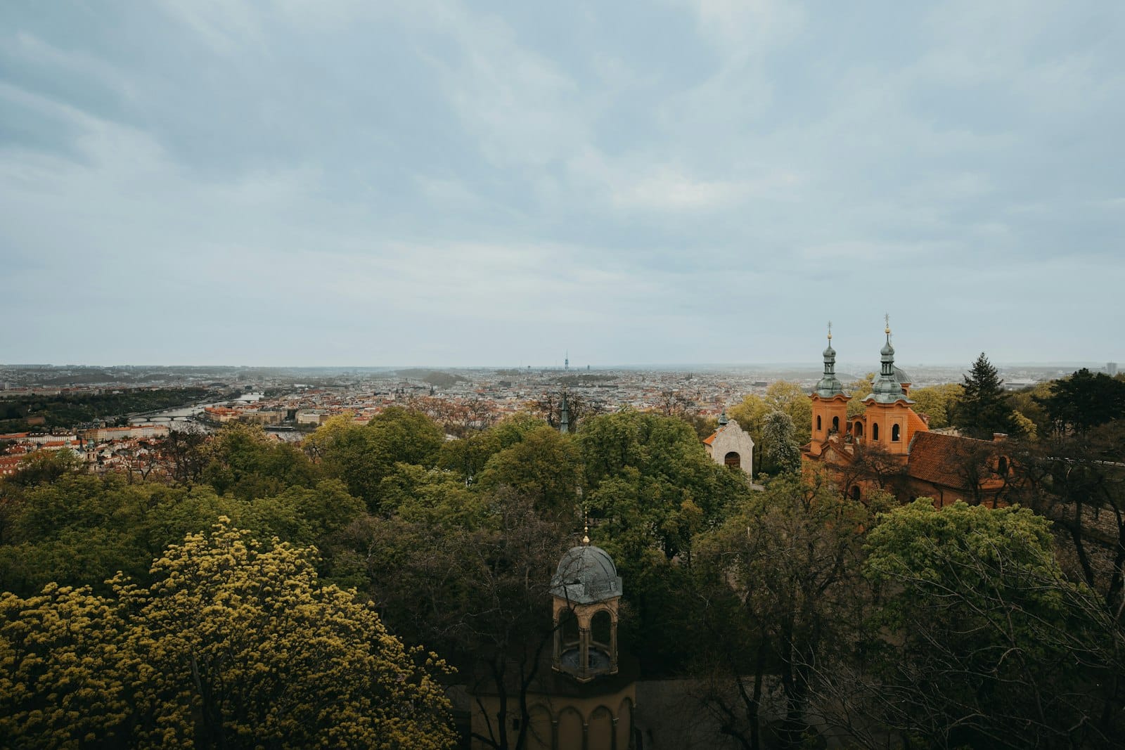 Weiter Blick vom Petřín-Hügel über Prag mit laubbedeckten Parkbäumen und der Kirche St. Michael im Vordergrund.