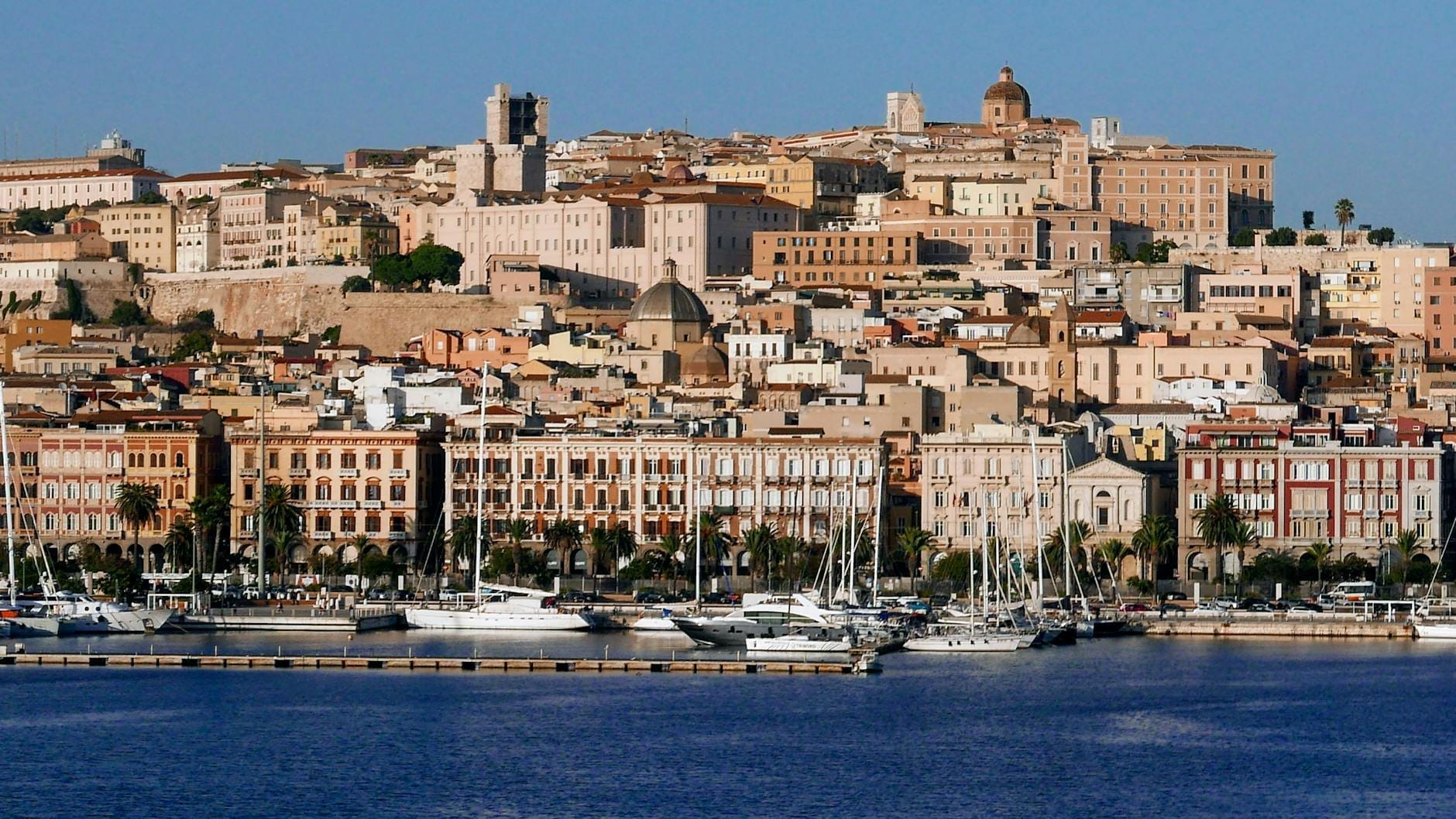 Panoramablick auf die Altstadt von Cagliari und den Yachthafen mit historischen Gebäuden und entlang der Uferpromenade angedockten Yachten unter einem klaren blauen Himmel.