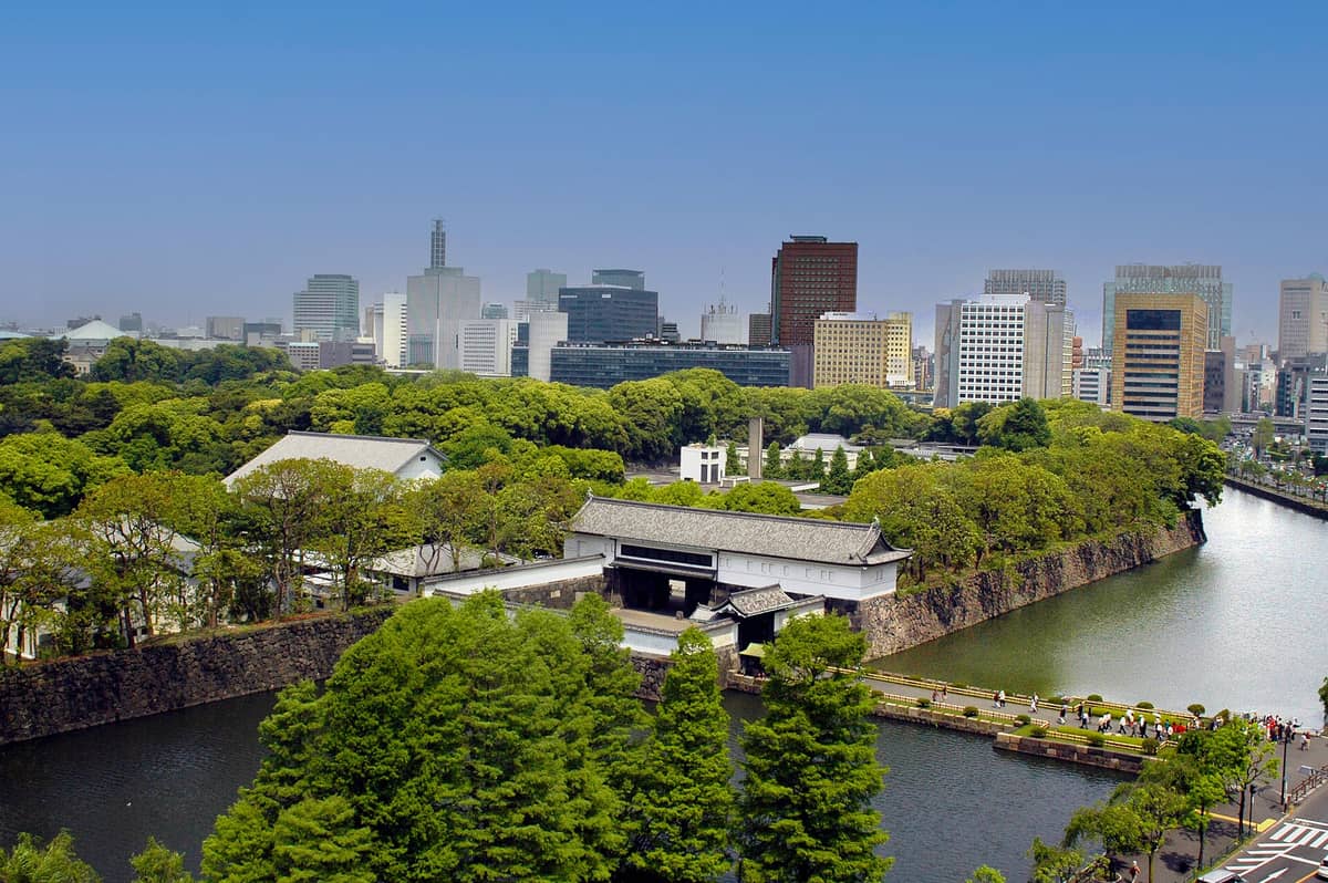Aerial view of Tokyo's Imperial Palace grounds with stone walls, moat, and traditional gate, city skyscrapers rising behind lush trees.