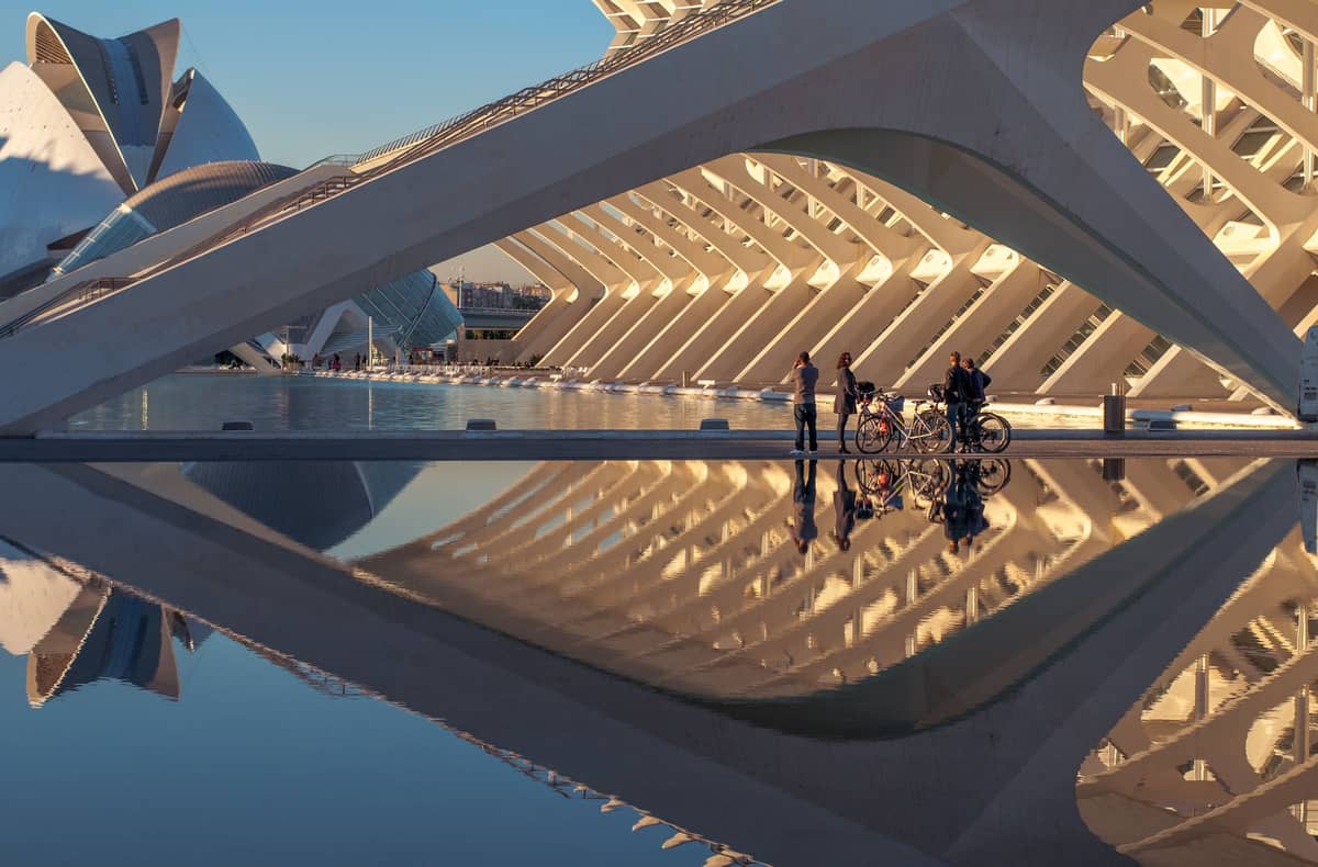 Moderne weiße Architekturstrukturen und breites Wasserbecken in der Ciudad de las Artes y las Ciencias in Valencia, mit im Wasser gespiegelten Radfahrern.