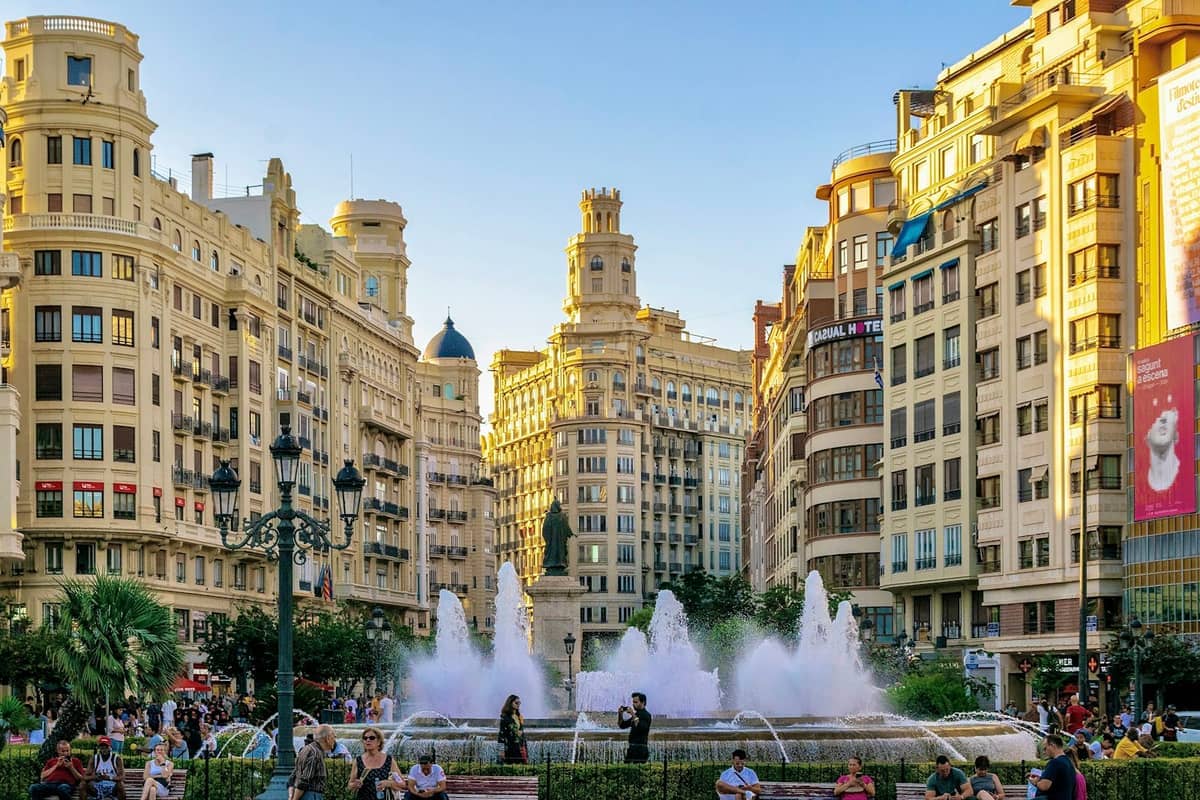 Plaza del Ayuntamiento a Valencia con edifici storici, una fontana centrale e persone che si rilassano sotto cieli azzurri nella luce del tardo pomeriggio.