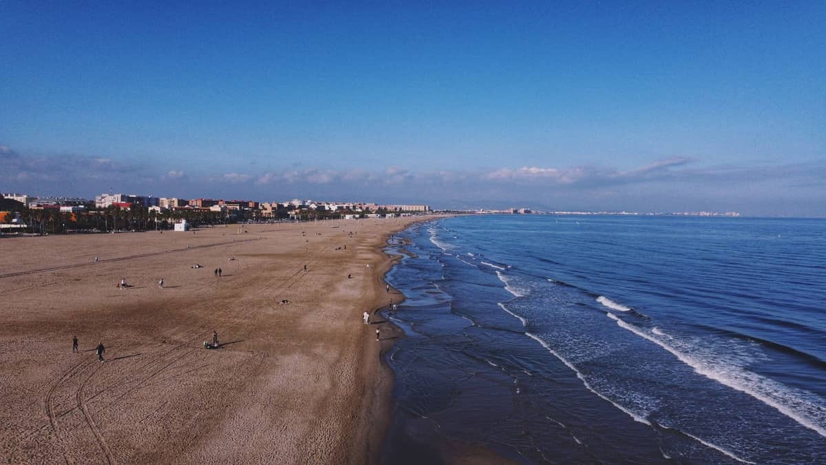 Weiter Sandstrand in Valencia mit sanften Wellen, fernen Stadtgebäuden und Menschen, die unter einem blauen Himmel am Ufer entlanggehen.