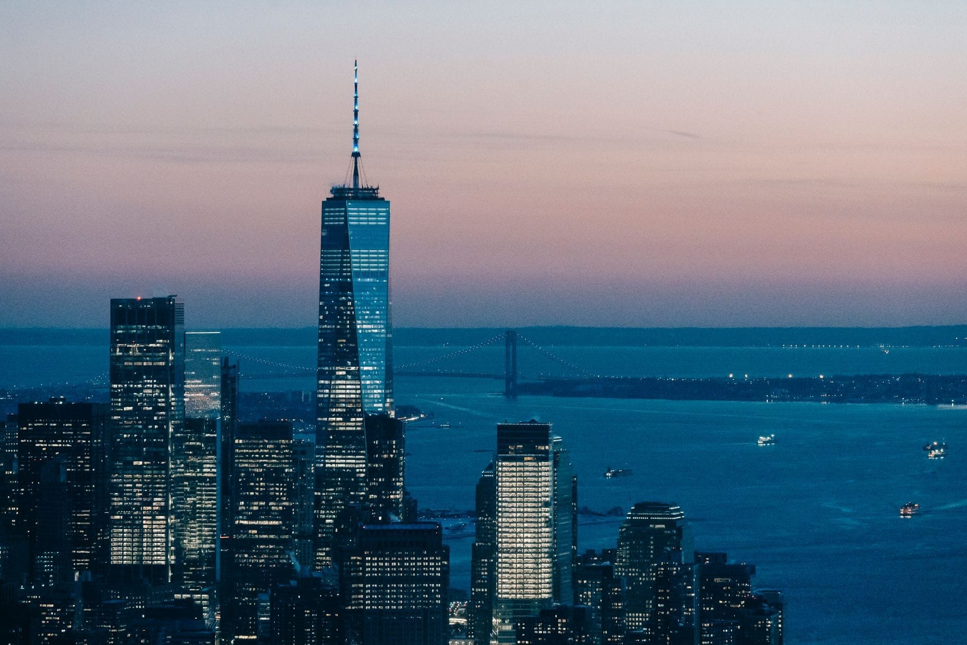 Vista del One World Trade Center che svetta sui grattacieli di Manhattan al crepuscolo, con l'Hudson River e un ponte lontano visibili.
