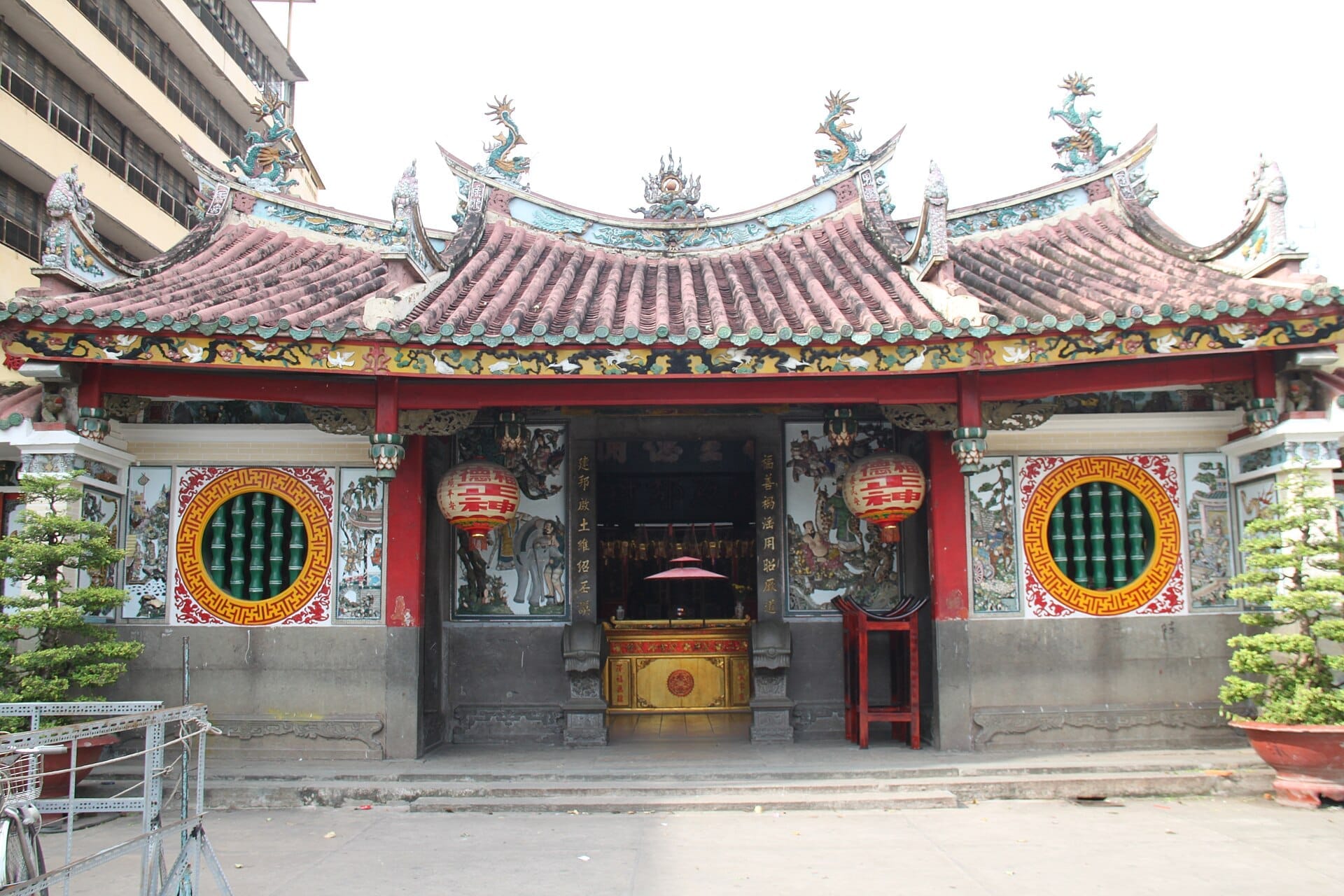 Vista frontal de la Pagoda Ông Bổn con tejado de azulejos ornamentados, murales coloridos, faroles rojos, ventanas circulares y plantas en macetas a la luz del día.