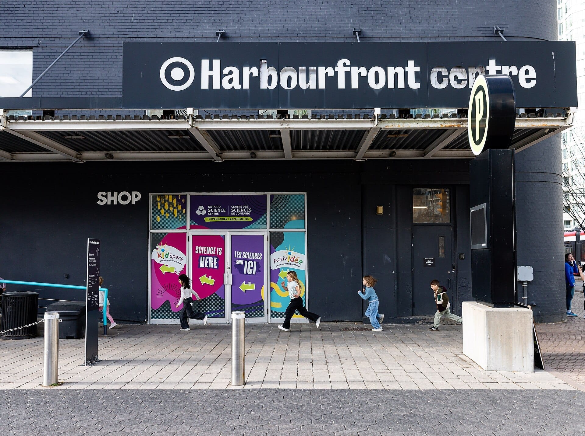 ทางเข้า Harbourfront Centre ที่มีป้าย Ontario Science Centre KidSpark พร้อมเด็กๆ วิ่งบนทางเท้าหน้าอาคารสีเข้ม