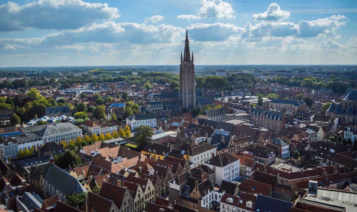 Aerial view over Bruges rooftops with the towering brick spire of the Church of Our Lady rising prominently in the center, surrounded by the historic cityscape.