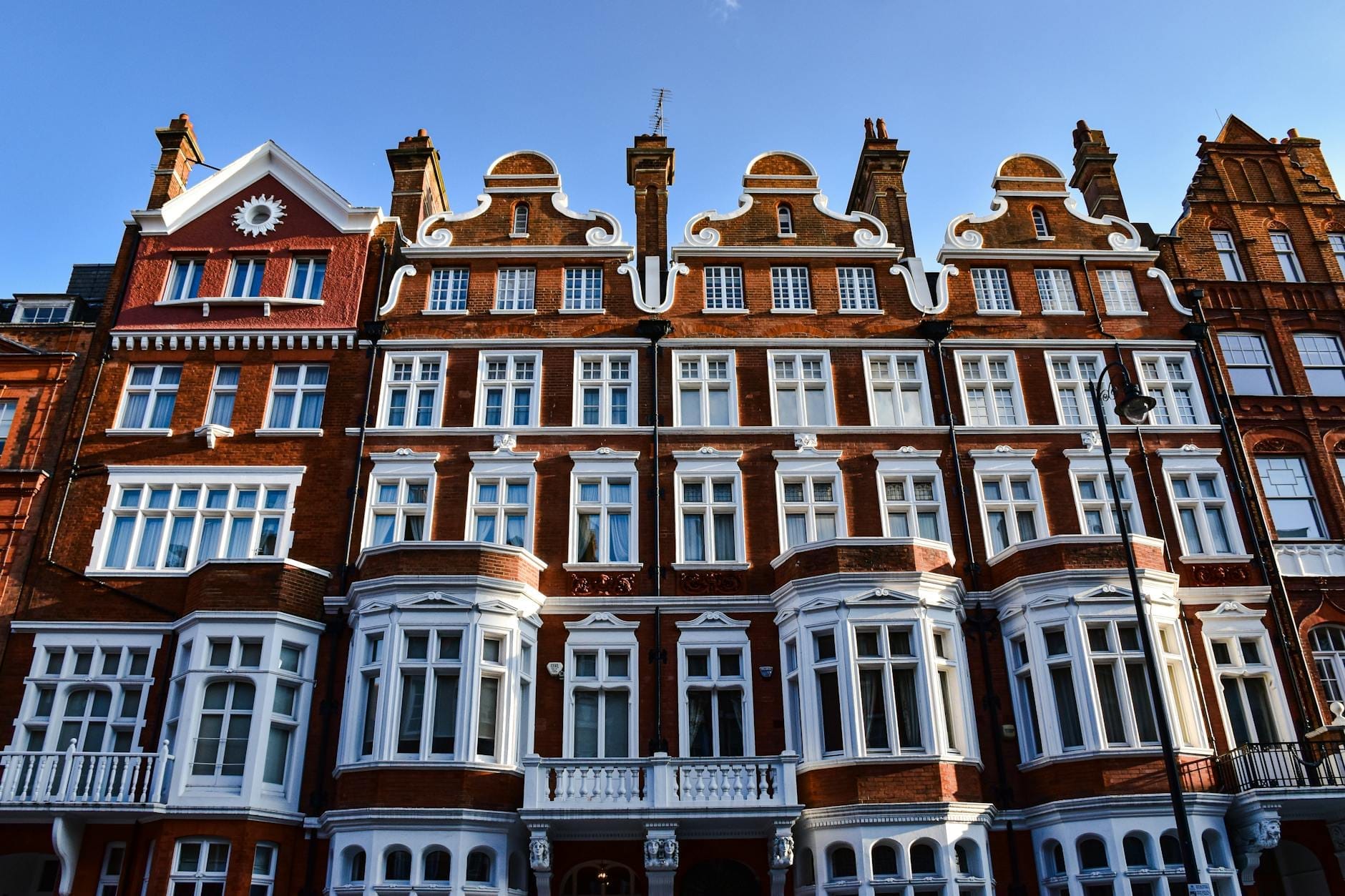 Uma fileira imponente de edifícios edwardianos ornamentados em tijolo vermelho com detalhes brancos sob um céu azul límpido, exibindo a arquitetura característica de Londres.