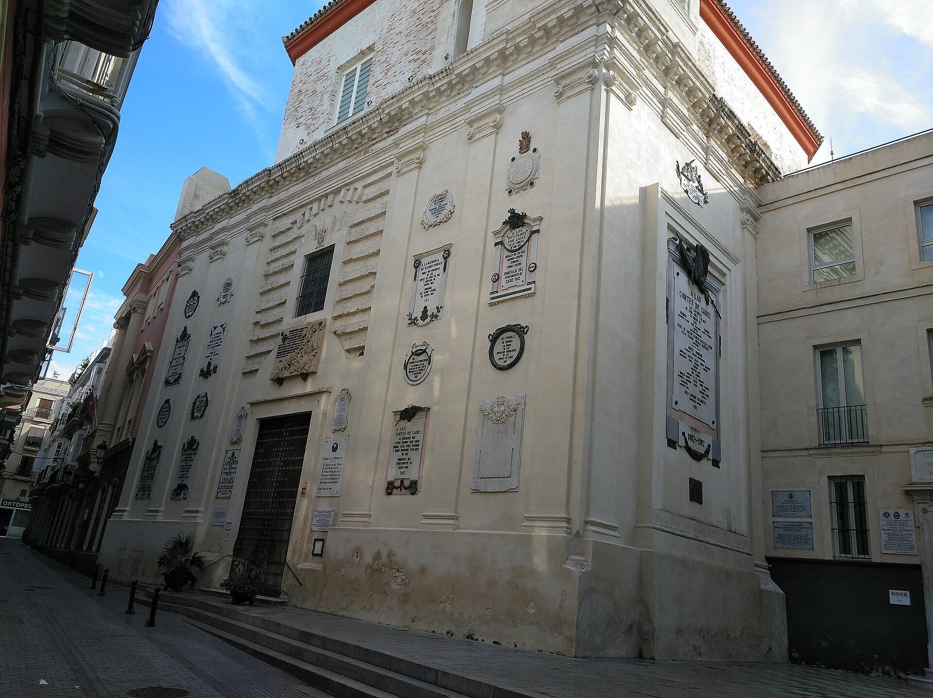 Facciata dell'Oratorio de San Felipe Neri a Cádiz, con pareti barocche color crema decorate con numerose targhe commemorative sotto un cielo azzurro sereno.