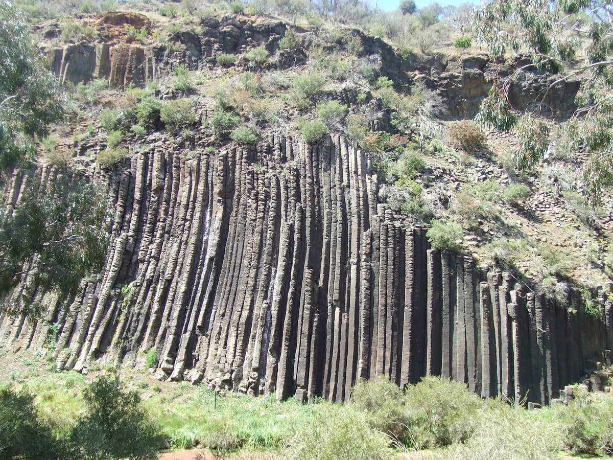 Vista de las icónicas formaciones de columnas de basalto conocidas como los Organ Pipes en el Parque Nacional Organ Pipes, rodeadas de vegetación y terreno escarpado.