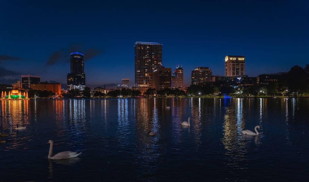 Lake Eola in der Dämmerung mit der Skyline von Orlando, Schwanenbooten im Vordergrund und bunten Lichtern, die sich im Wasser spiegeln.