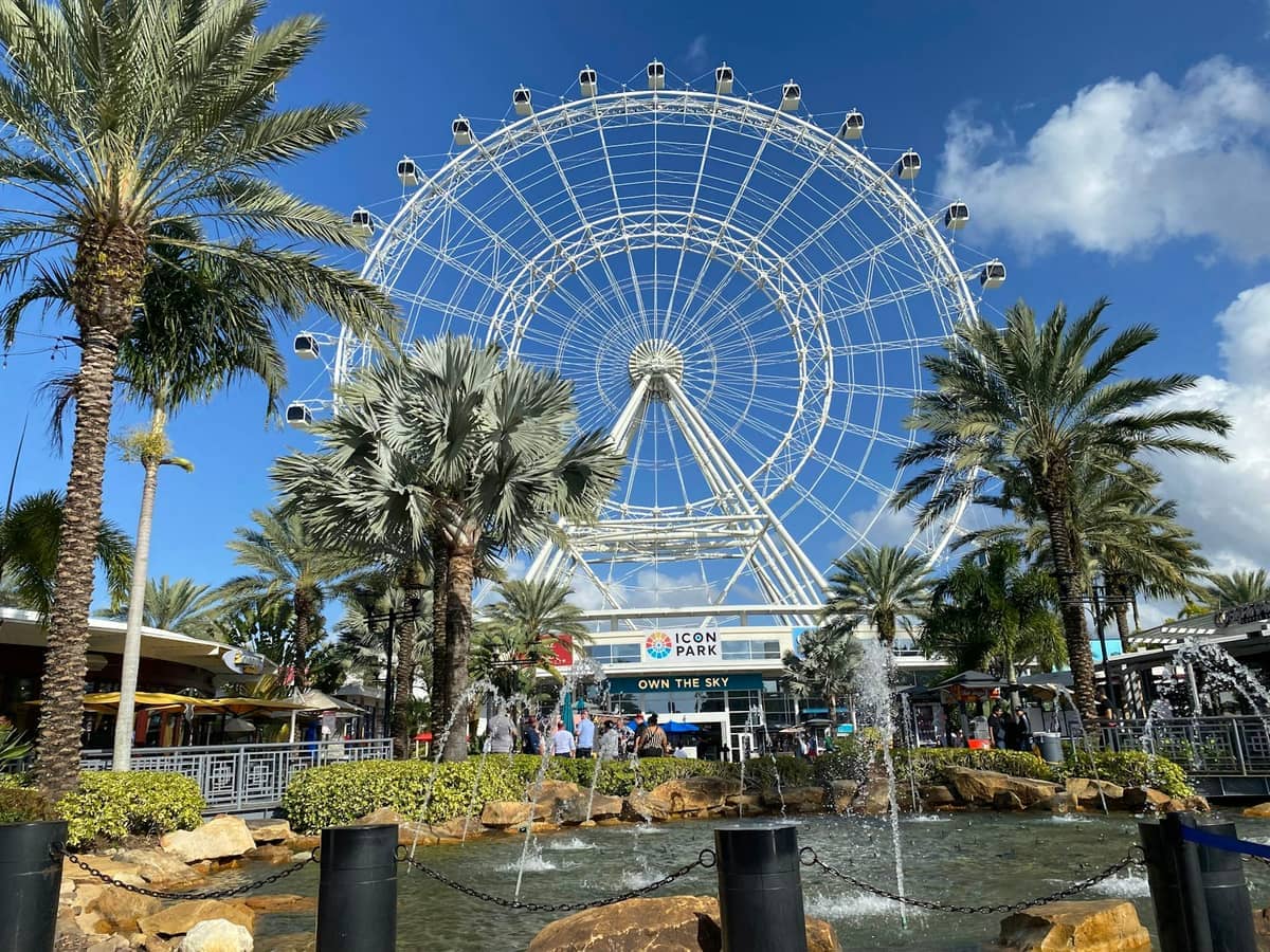 The Orlando Eye Ferris wheel at ICON Park towers above palm trees, fountains, and visitors under a sunny blue sky with scattered clouds.