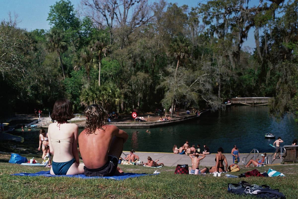 Menschen entspannen sich an einem grasbewachsenen Hang mit Blick auf einen natürlichen Quell-Badebereich, umgeben von üppigen Bäumen und Schwimmern im Wasser.