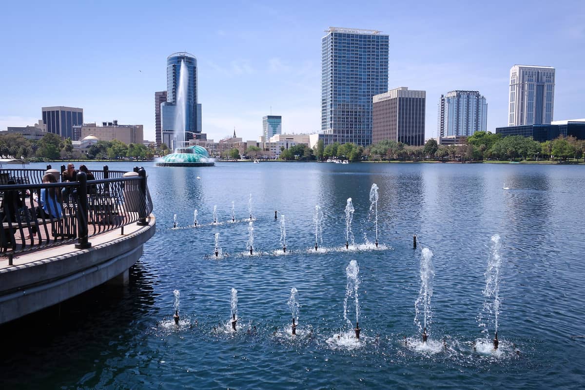 Sonnige Aussicht auf den Lake Eola Park in Orlando, Florida, mit Menschen auf einem Uferweg, kleinen Fontänen im Wasser und der Skyline der Innenstadt im Hintergrund.