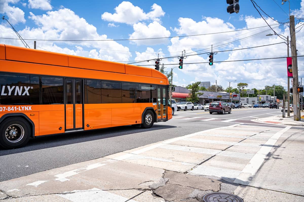 Un autobus pubblico arancione LYNX in movimento a un incrocio soleggiato di Orlando con traffico, linee elettriche e cielo blu nuvoloso.