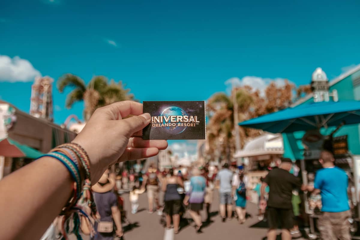 Person holding a Universal Orlando Resort ticket in front of a busy street scene at Universal Orlando, with palm trees, park buildings, and a sunny blue sky.