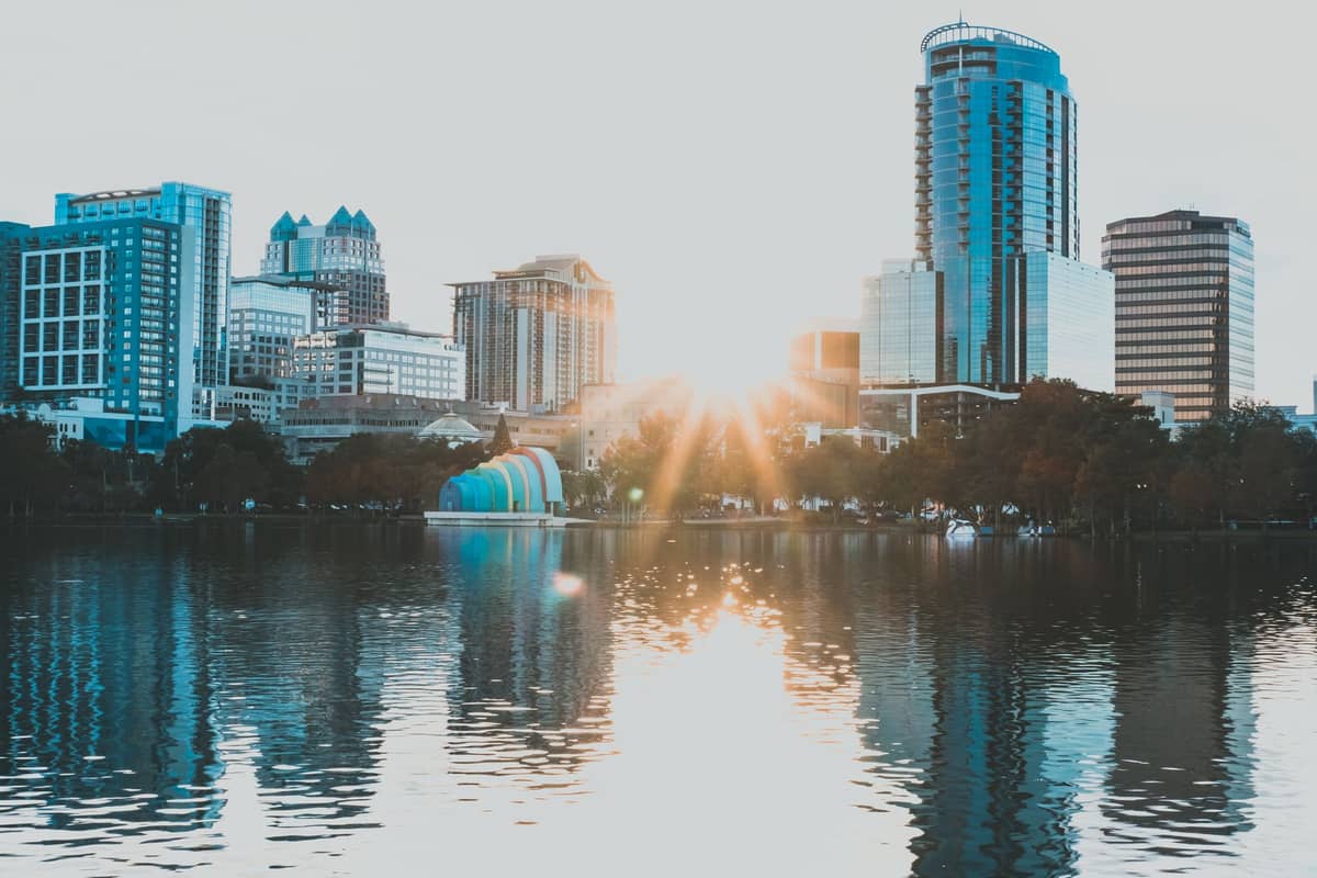 Skyline de Orlando con modernos rascacielos reflejados en el lago Eola al amanecer, con árboles bordeando la orilla en primer plano.