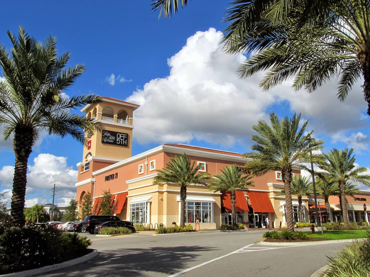 Orlando Vineland Premium Outlets bei hellem Tageslicht mit Palmen, Parkplätzen und Ladenfronten unter einem lebhaften blauen Himmel mit vereinzelten Wolken.