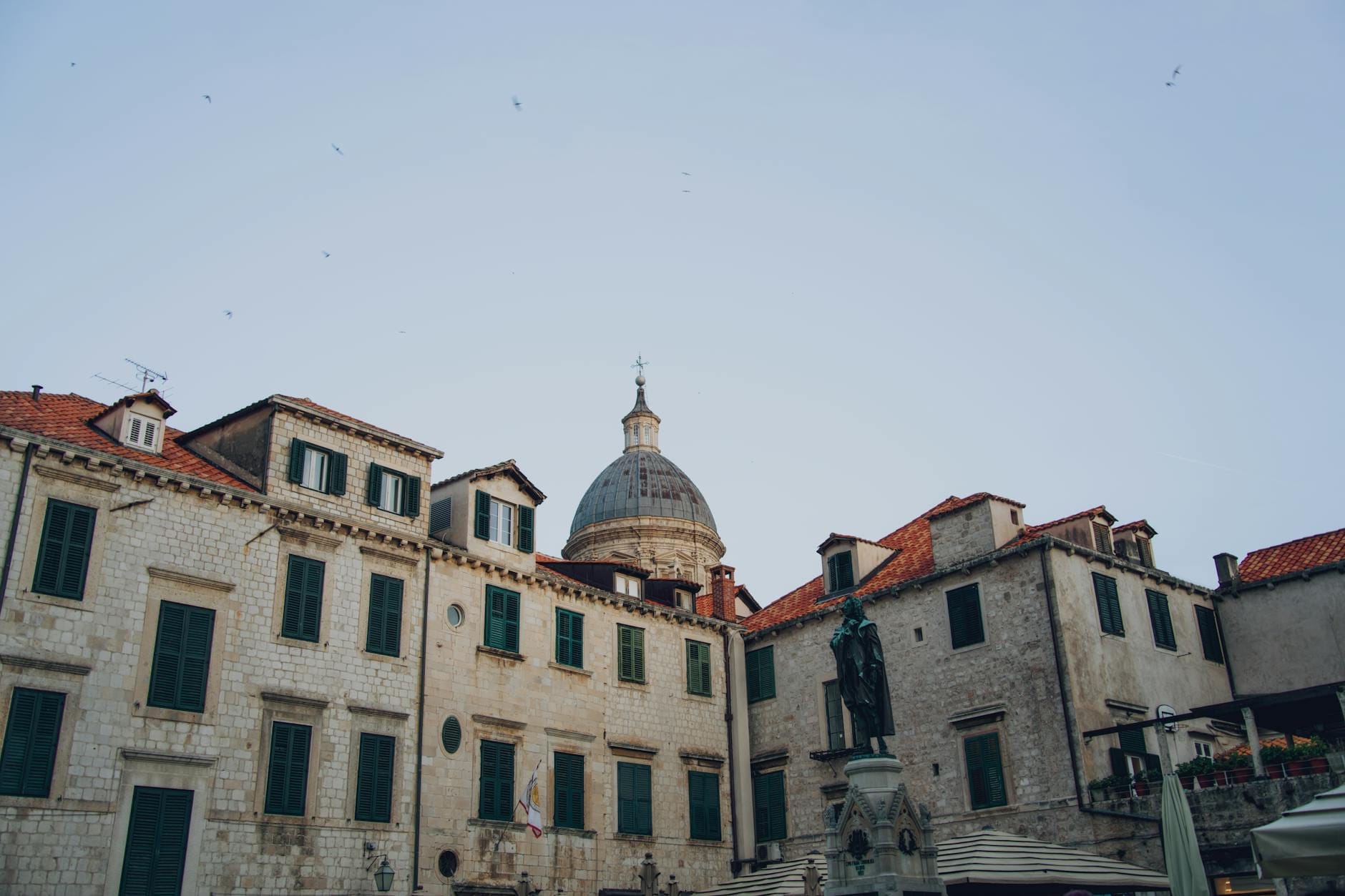 Vista de la plaza Luža en el casco antiguo de Dubrovnik con edificios históricos de piedra caliza, una iglesia con cúpula destacada y la Columna de Orlando visible en el centro.
