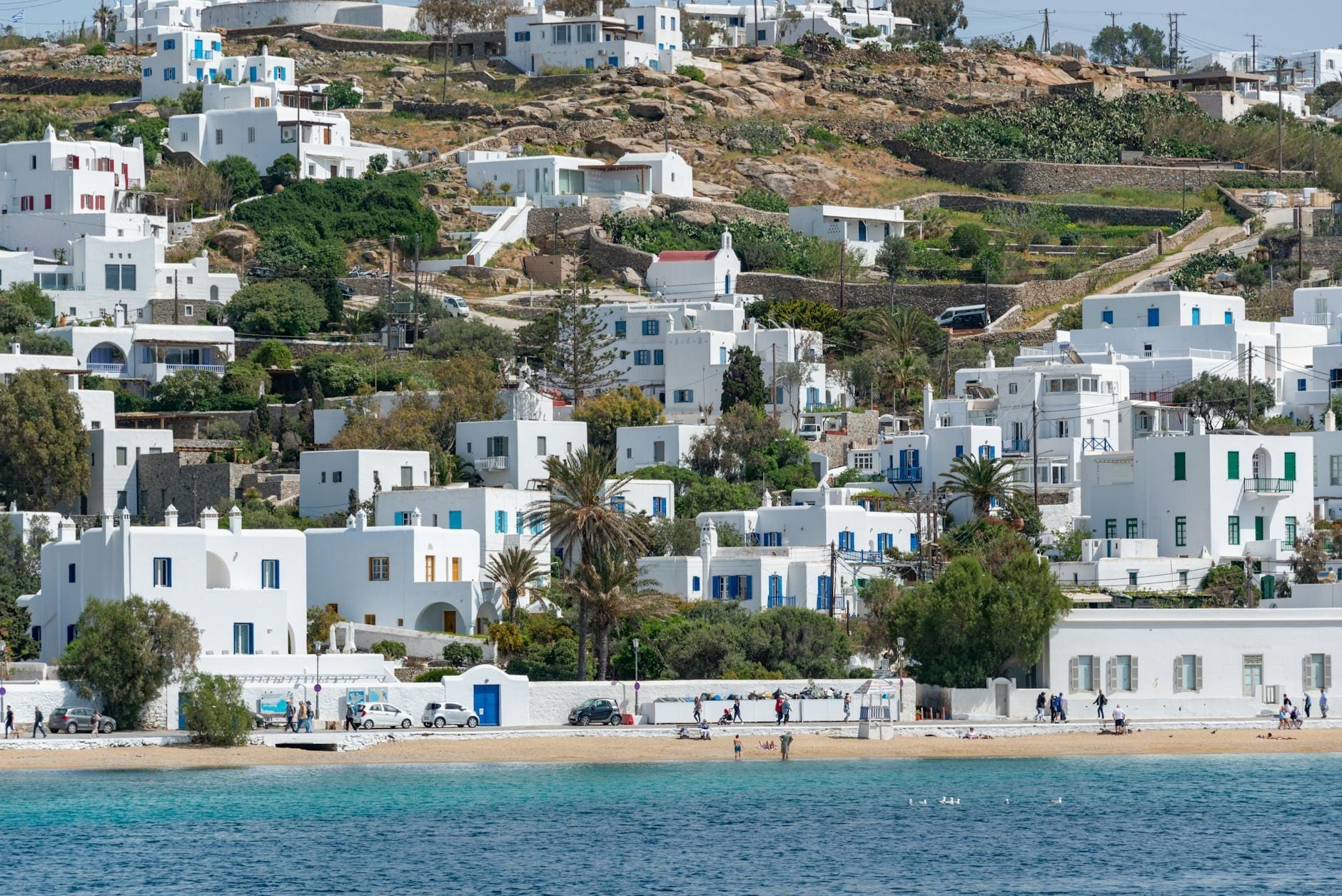 Vista panorámica de la playa de Ornos con gente en la orilla arenosa, aguas azules y cristalinas, y racimos de casas cicládicas blancas que trepan por la ladera en Mykonos.