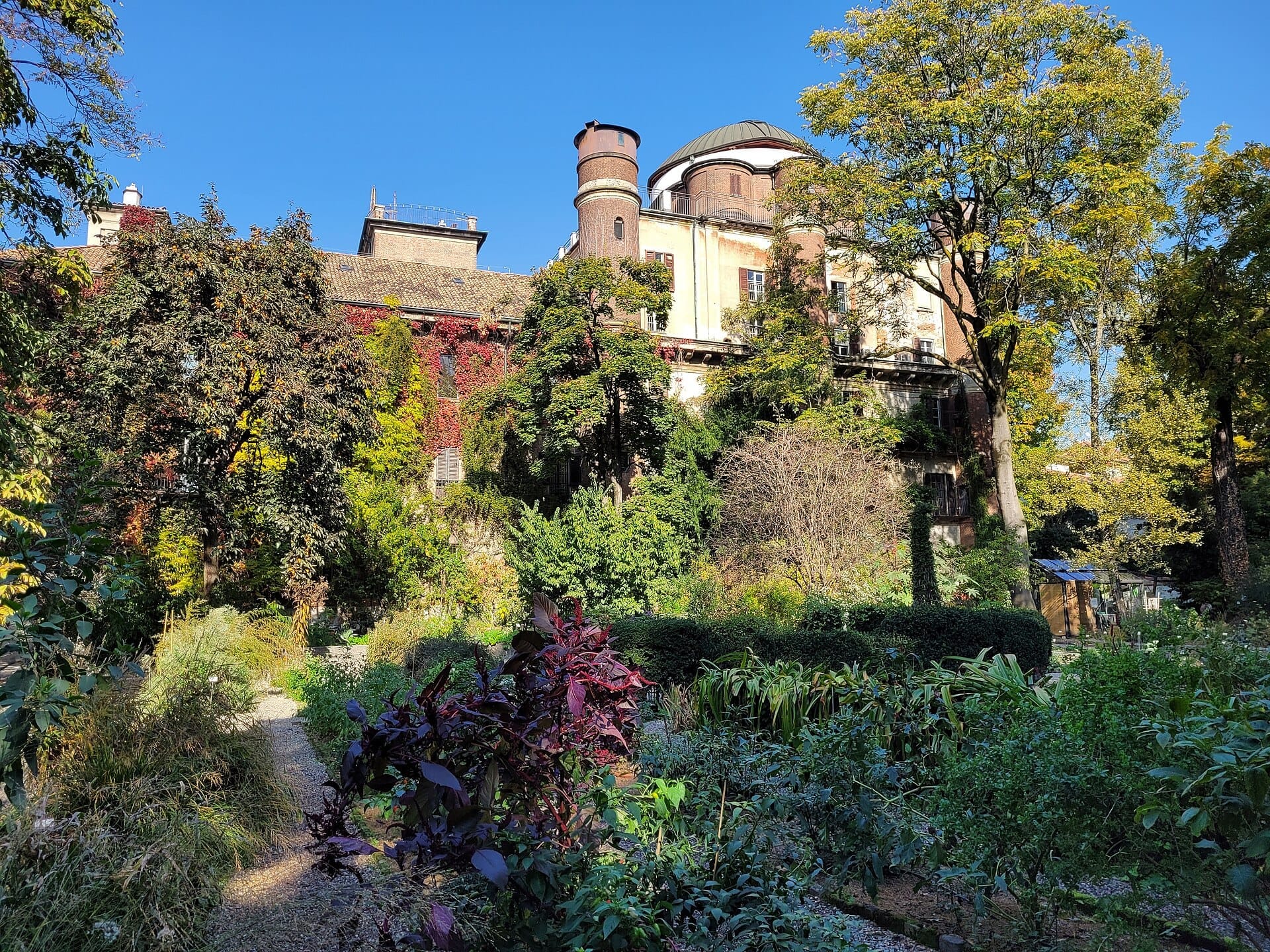 Vista soleggiata di rigogliosi giardini, alberi maturi e gli storici edifici del Palazzo Brera nell'Orto Botanico di Brera, Milano.