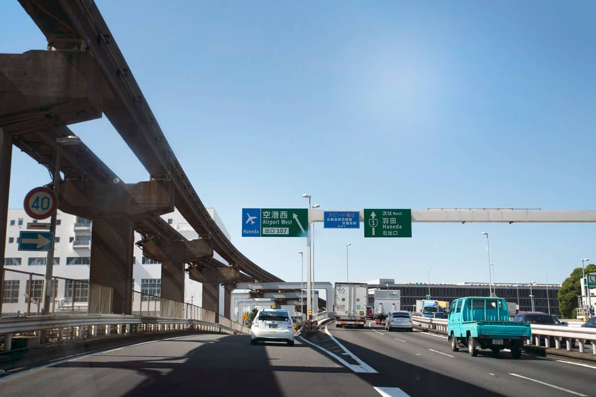 Highway and monorail tracks leading toward Itami Airport, with green road signs indicating airport directions and cars driving below a blue sky.