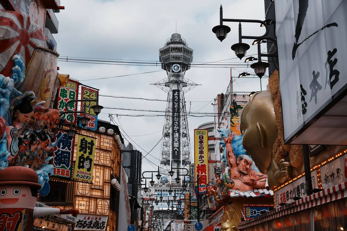 Bustling Osaka street scene with Tsutenkaku Tower in the background, colorful neon signs, and some character-themed shop displays visible.