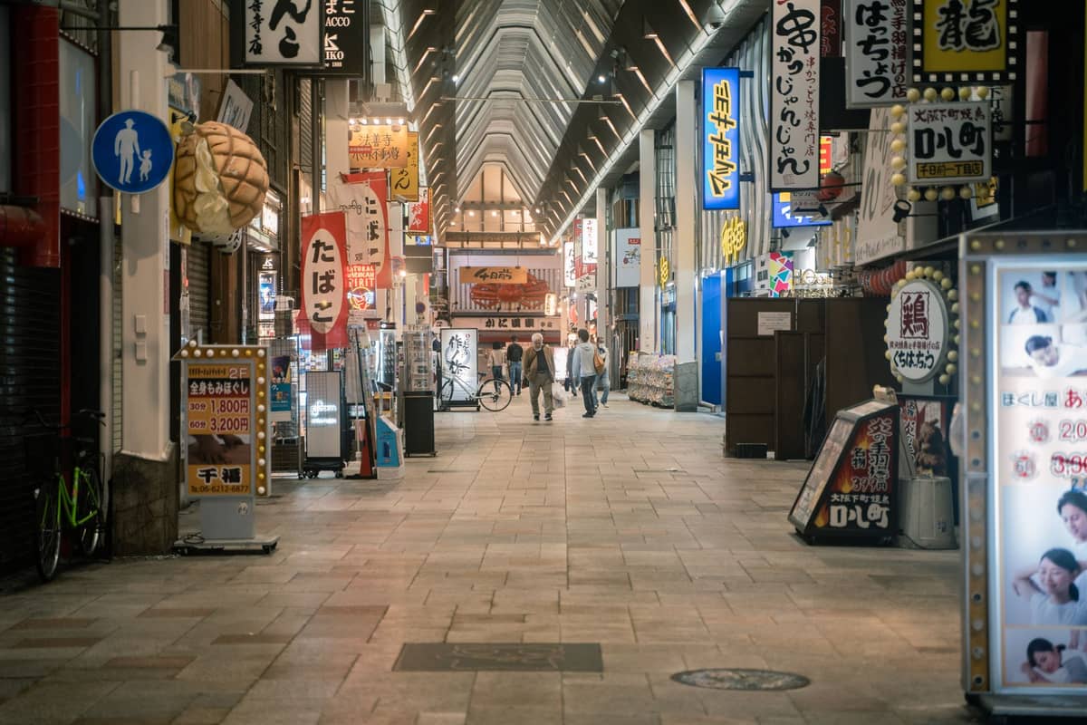 A covered shopping arcade in Den Den Town, Osaka, with anime and game store signs, bright lights, and shoppers walking along the pedestrian street.