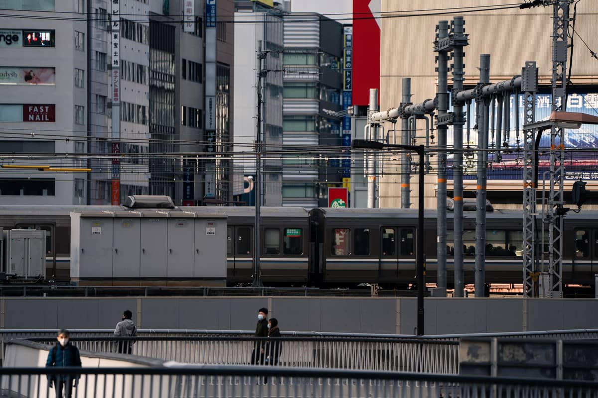 Urban Osaka scene with train tracks, a commuter train, and pedestrians on a walkway surrounded by city buildings.