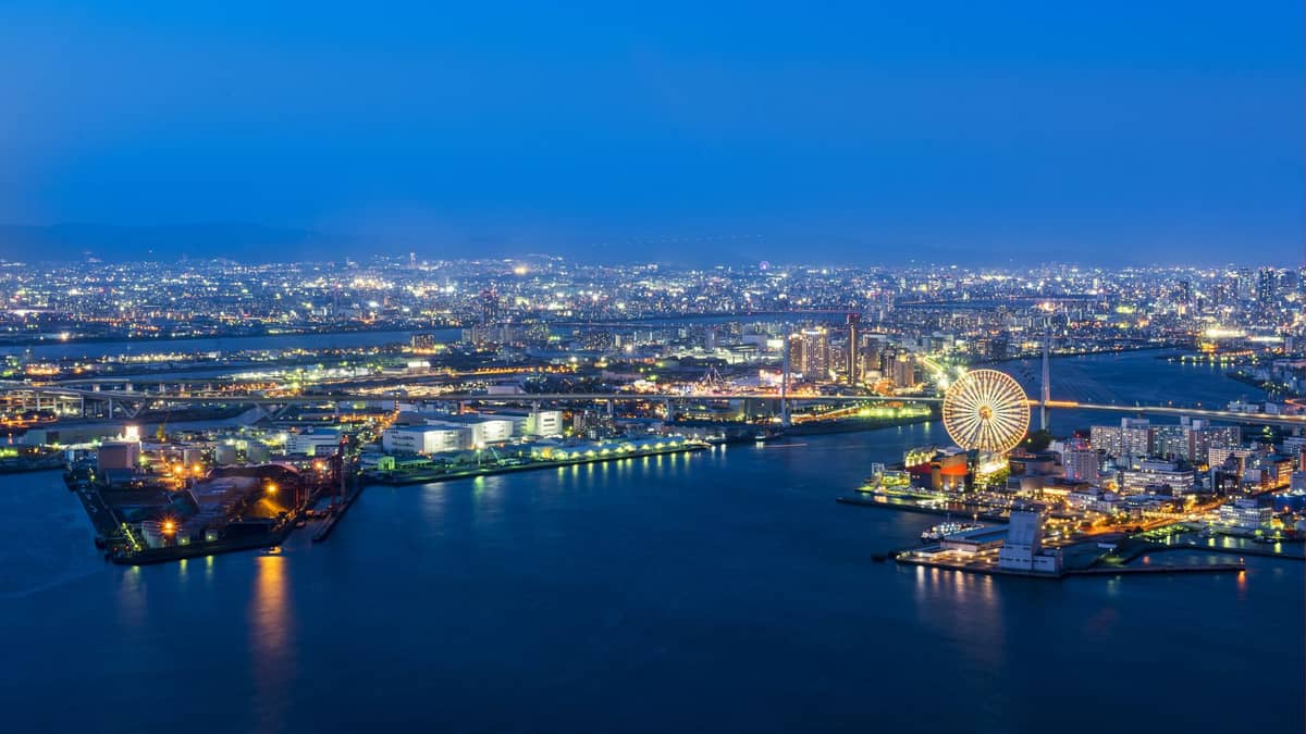 Aerial evening view of Osaka Bay Area with the waterfront, illuminated ferris wheel, and leisure attractions along the coastline under a deep blue sky.