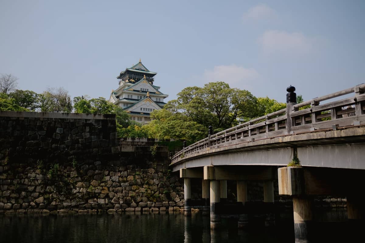 Osaka Castle viewed from across the moat and bridge, surrounded by trees and stone walls under a blue sky.