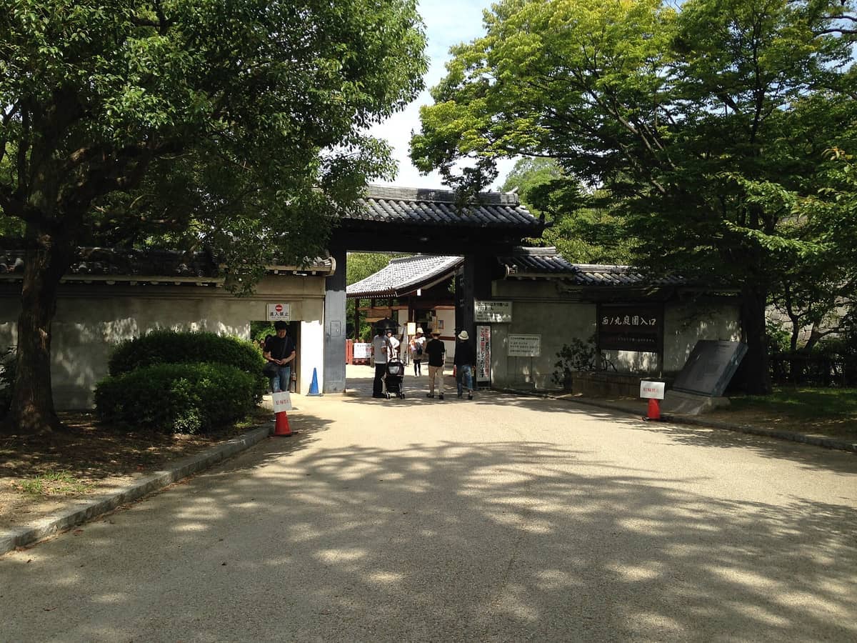 Entrance to Nishinomaru Garden at Osaka Castle, framed by traditional stone walls and lush green trees on a sunny day.