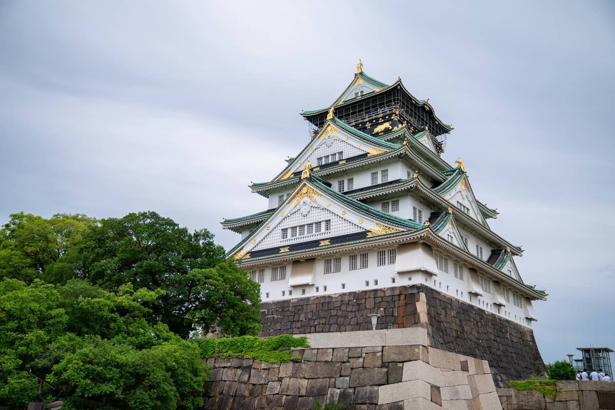 Osaka Castle stands on a stone foundation surrounded by lush green trees, its white walls and gold accents under an overcast sky.